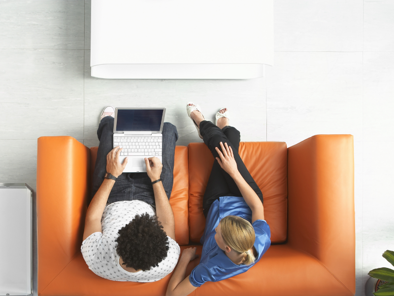 Overhead view of a man and woman looking at a laptop computer while sitting on an orange loveseat.